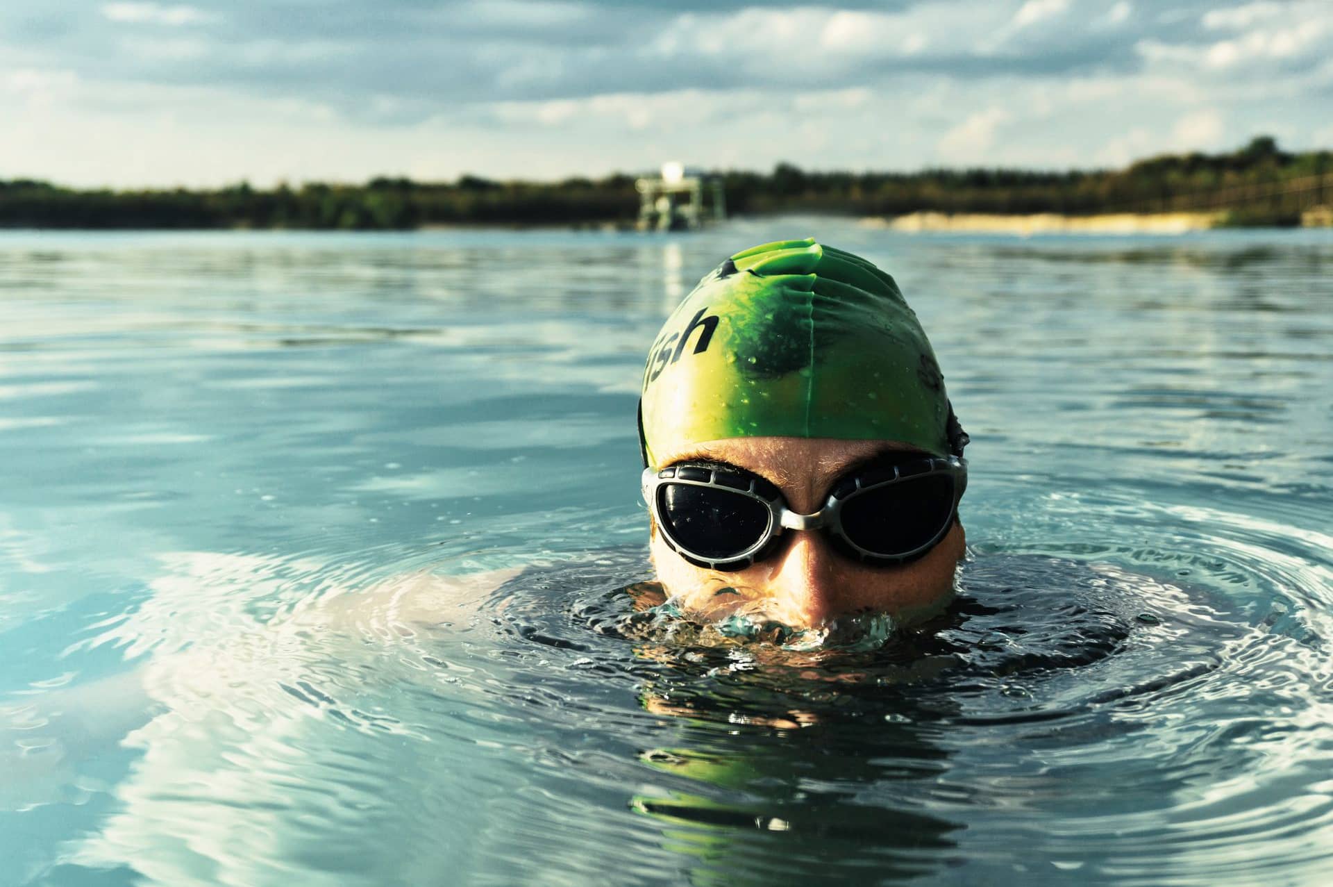 A swimmer in a cap and goggles coming out of a calm lake on a sunny day